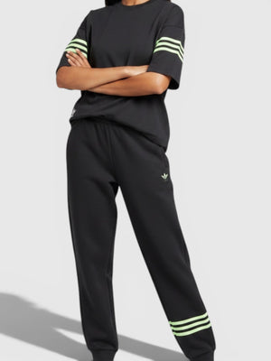 Model wanita mengenakan setelan Adidas Neuclassics hitam dengan aksen 3-stripes hijau neon dan potongan loose fit