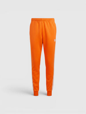 Celana track Adidas Adicolor Classics SST berwarna oranye dengan detail 3-Stripes putih dan logo Trefoil dari FTFO || BIG BRAND LOW PRICE