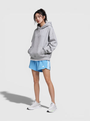 Model mengenakan celana pendek olahraga Adidas Pacer WVN High biru dengan detail 3-stripes putih dan hoodie abu-abu