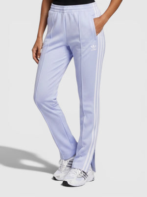Model wanita mengenakan celana track Adidas Adicolor Classics SST berwarna ungu muda dengan detail 3-Stripes putih