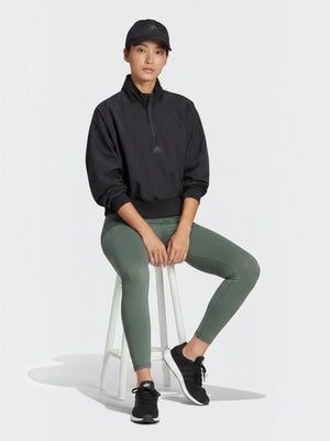 Model mengenakan jaket Adidas Z.N.E. WVN QZ hitam dengan quarter-zip dan celana legging hijau saat duduk di kursi.