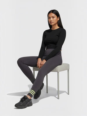 Model mengenakan legging Adidas Adicolor Neuclassics abu-abu dengan detail 3-Stripes kuning neon di pergelangan kaki
