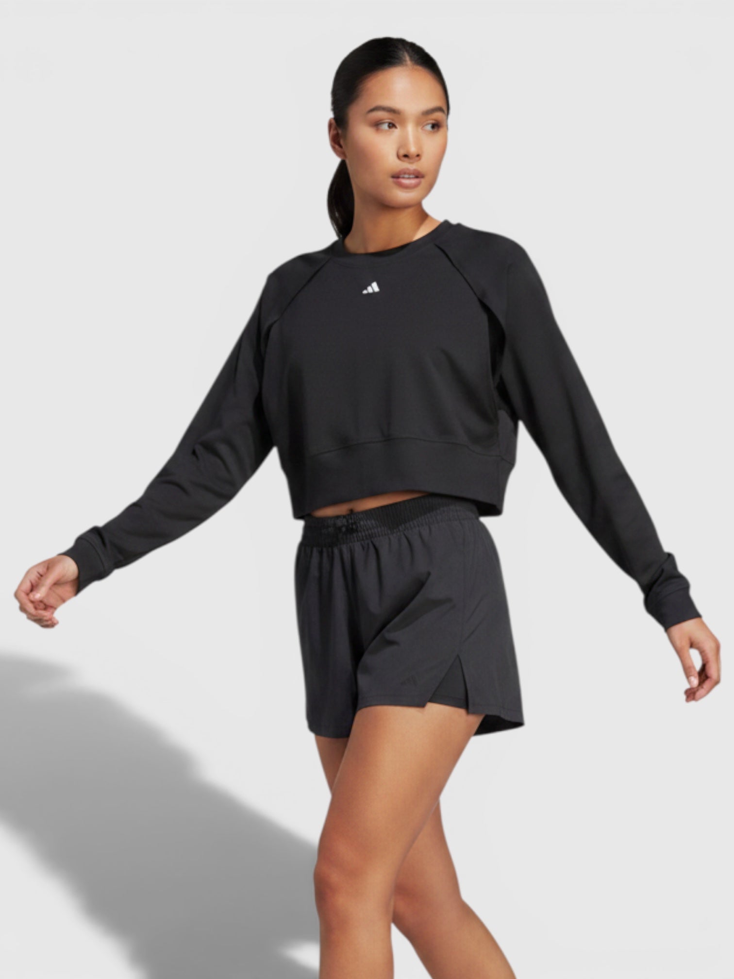 Model mengenakan Adidas Power AEROREADY crop sweatshirt hitam dengan lengan unik dan celana pendek olahraga senada