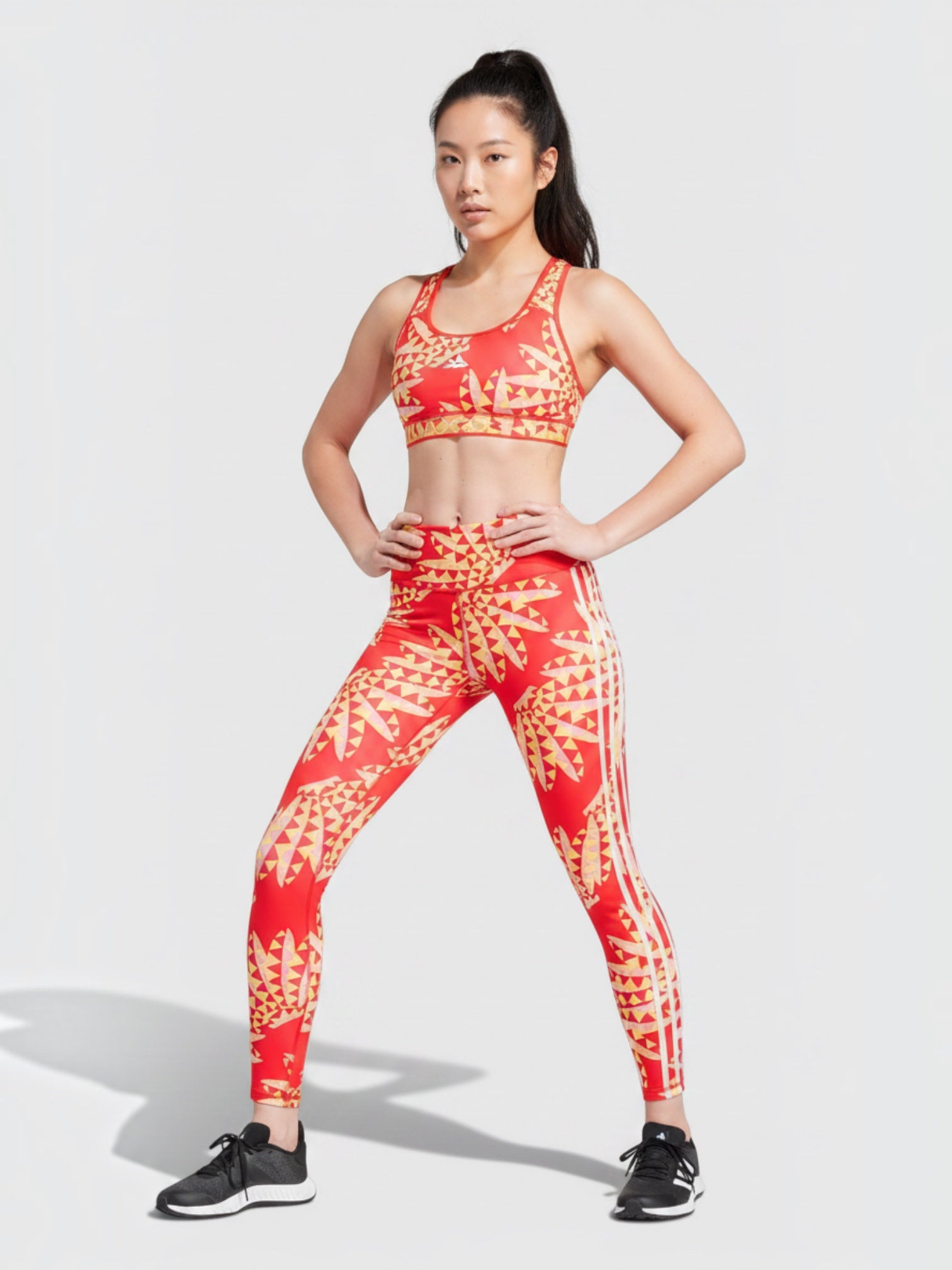 Model mengenakan set olahraga Adidas x FARM Rio berwarna merah dengan motif kaleidoskop tropis dan aksen tiga garis.