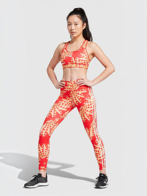 Model mengenakan set olahraga Adidas x FARM Rio berwarna merah dengan motif kaleidoskop tropis dan aksen tiga garis.