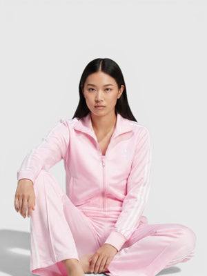 Model wanita mengenakan setelan track suit Adidas Firebird warna merah muda dengan detail 3-Stripes putih