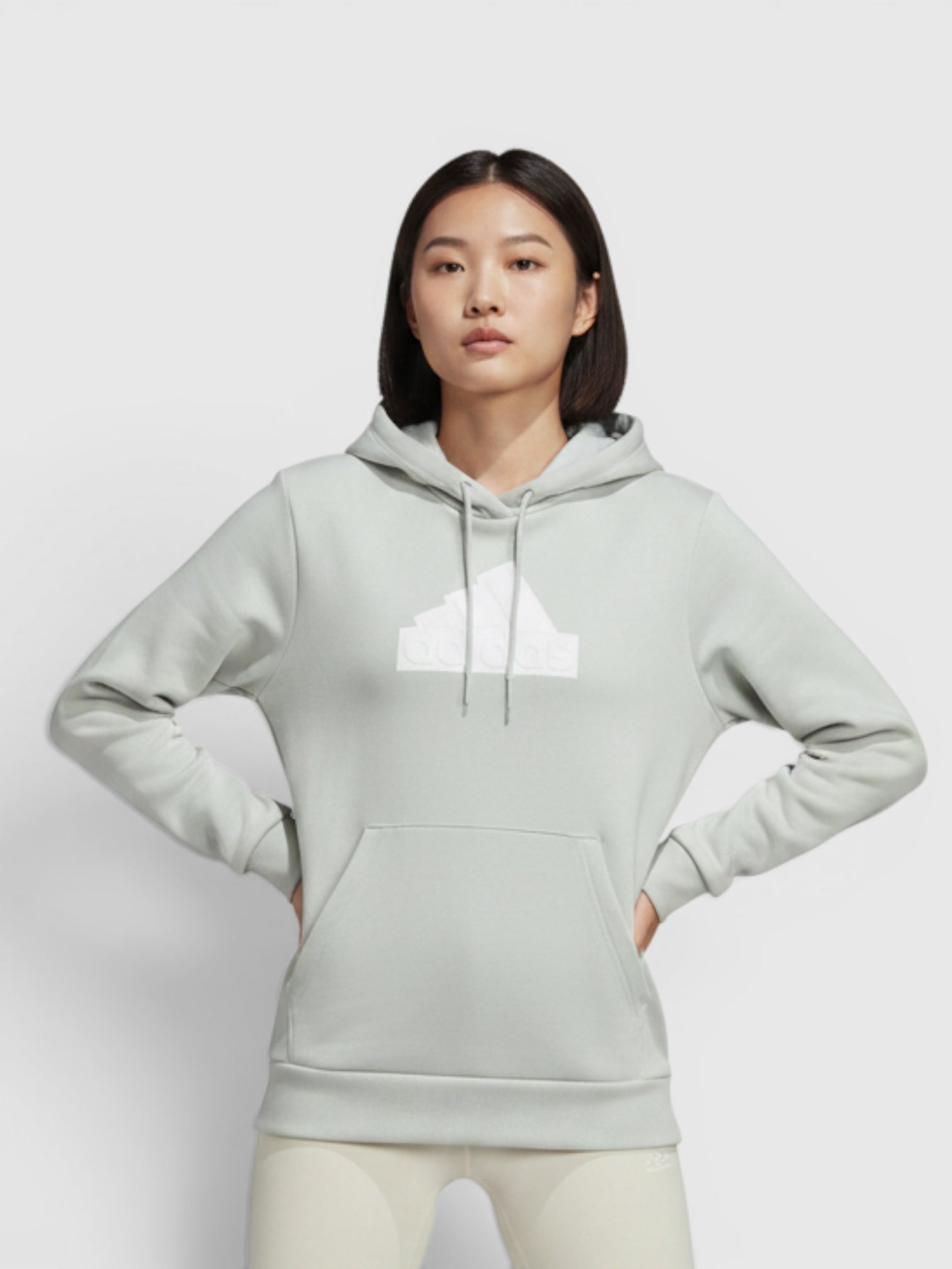 Model wanita mengenakan hoodie Adidas Future Icons Badge of Sport berwarna abu-abu muda dengan logo puff-print putih dari FTFO || BIG BRAND LOW PRICE