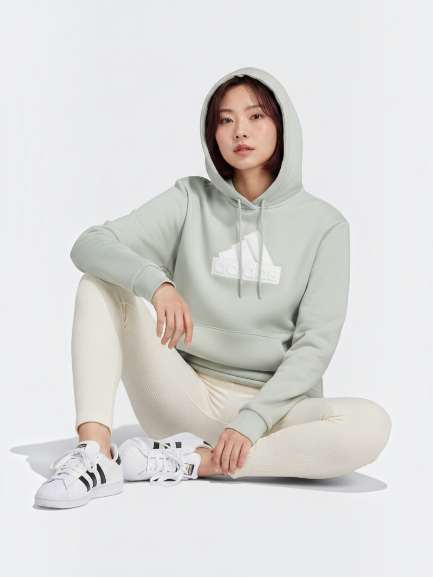 Model wanita mengenakan hoodie Adidas Future Icons berwarna hijau sage dengan logo Badge of Sport putih yang timbul.