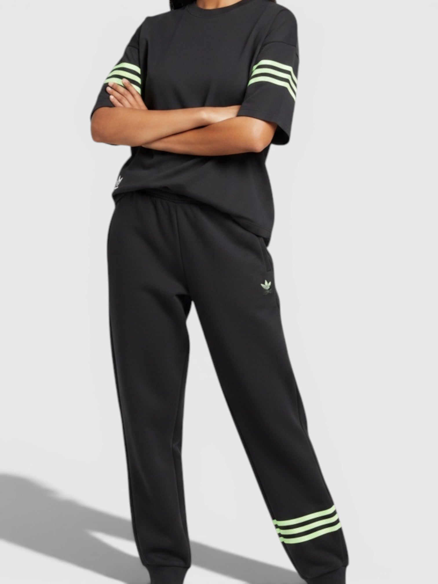 Model wanita mengenakan setelan Adidas Neuclassics hitam dengan aksen 3-stripes hijau neon dan potongan loose fit