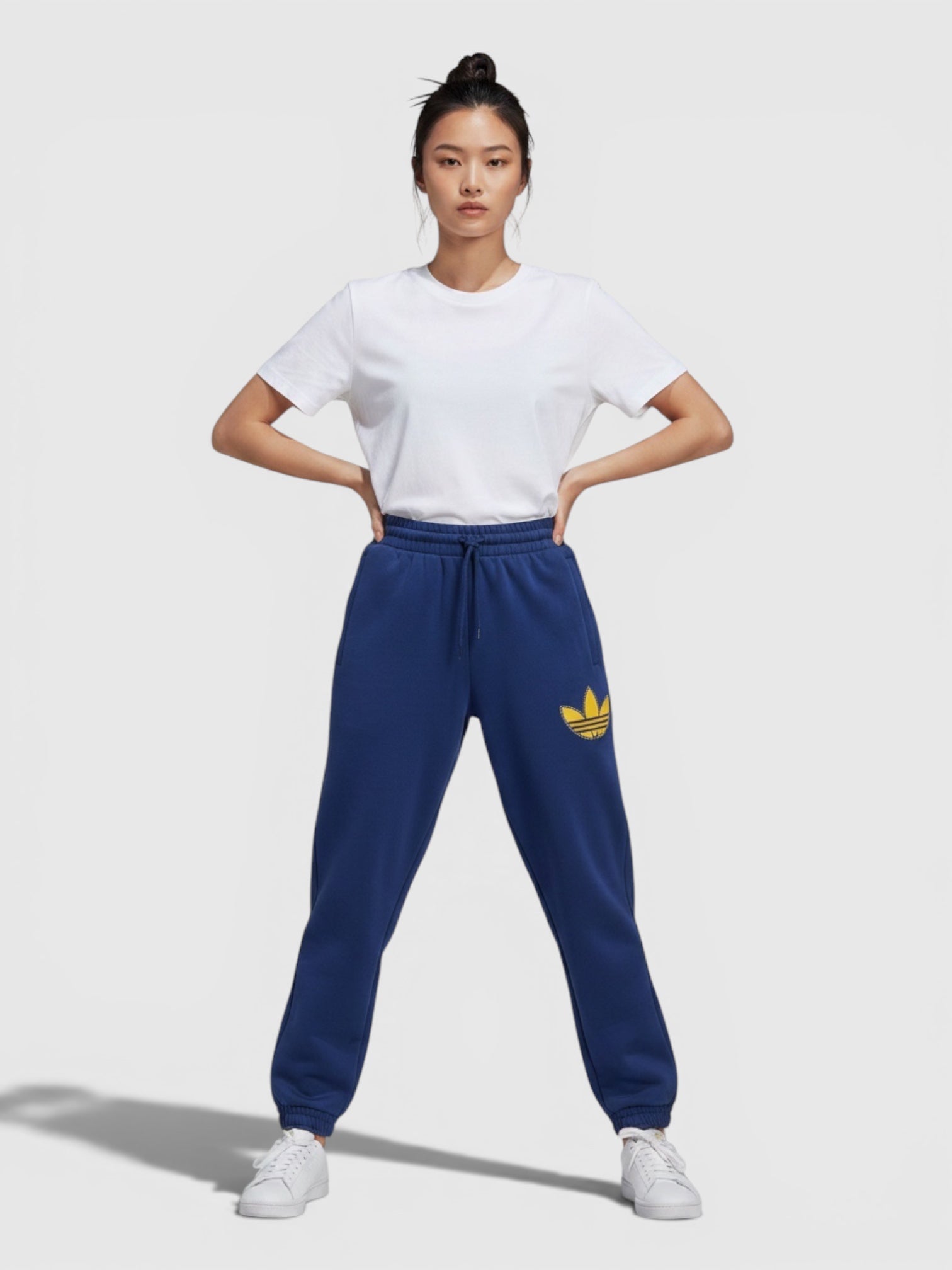 Model wanita mengenakan celana jogger Adidas biru dengan logo trefoil kuning dan detail cuffed di pergelangan kaki