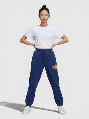 Model wanita mengenakan celana jogger Adidas biru dengan logo trefoil kuning dan detail cuffed di pergelangan kaki