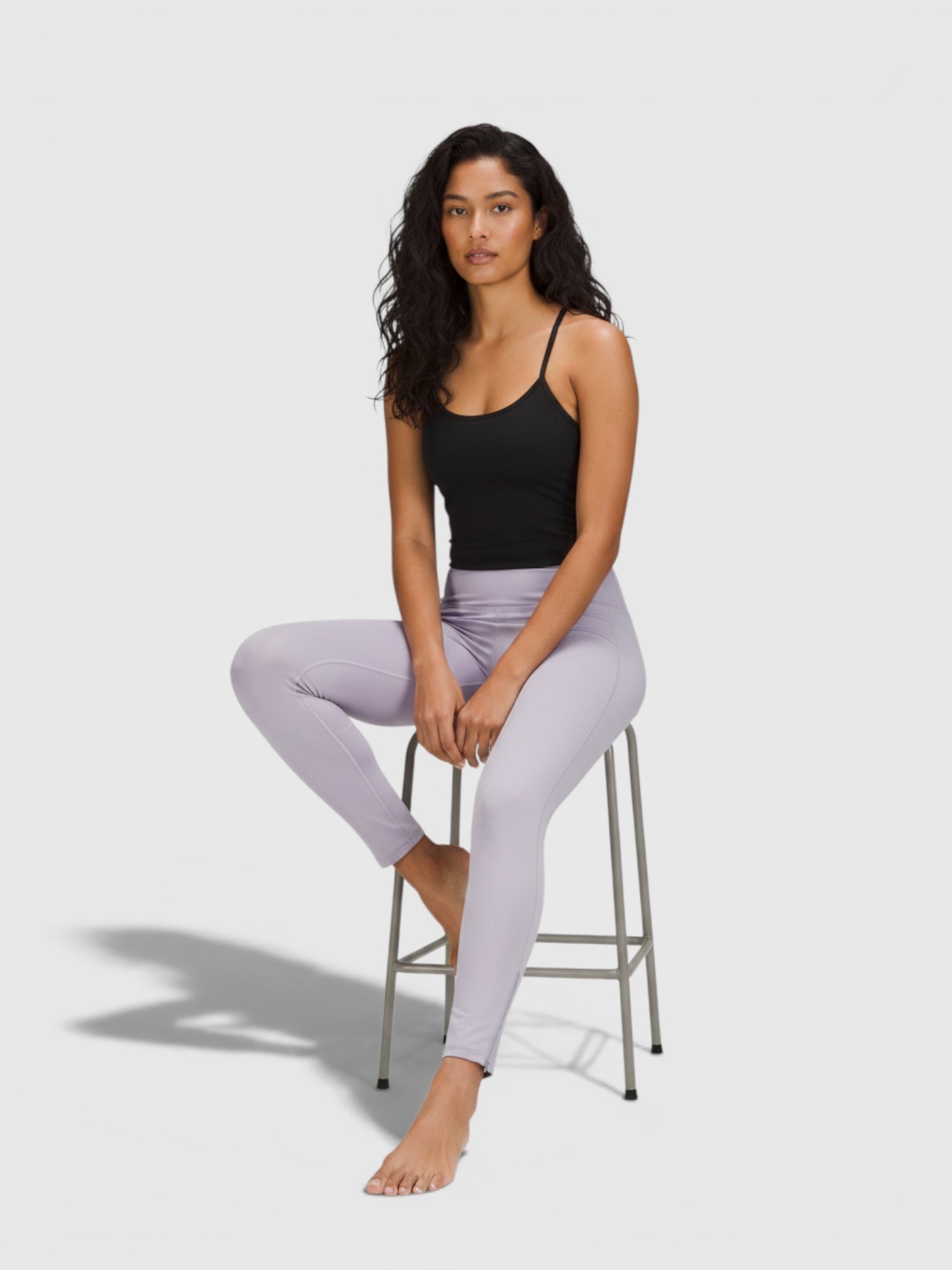 Model wanita mengenakan celana ketat Adidas W New 7/8 Tights berwarna ungu muda dengan atasan hitam saat duduk di kursi