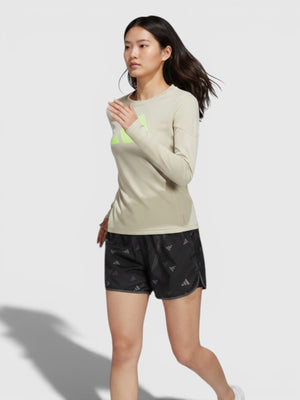 Model wanita mengenakan celana lari Adidas Run It Brand Love hitam dengan grafis logo dan atasan lengan panjang krem