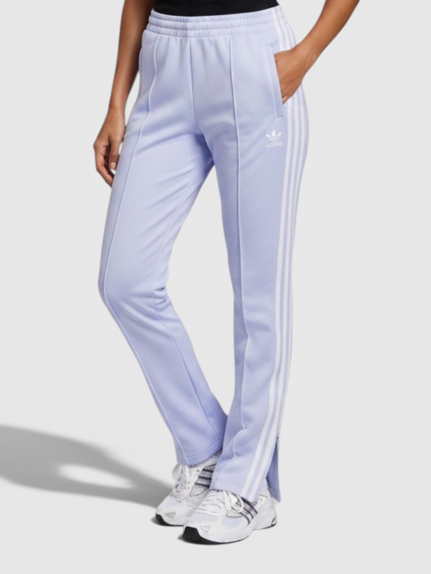 Model wanita mengenakan celana track Adidas Adicolor Classics SST berwarna ungu muda dengan detail 3-Stripes putih