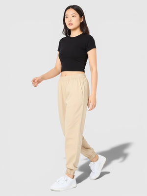 Model wanita mengenakan celana track Adidas Essentials+ berwarna krem dengan potongan tapered dan manset elastis