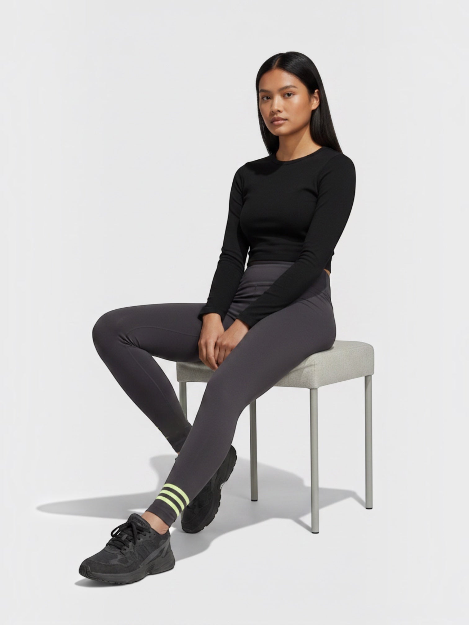 Model mengenakan legging Adidas Adicolor Neuclassics abu-abu dengan detail 3-Stripes kuning neon di pergelangan kaki